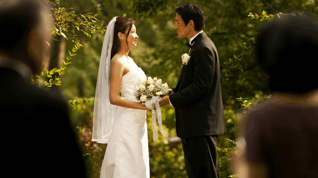 Um casal composto por uma mulher e um homem estão casando a luz do dia. Eles estão parados um de frente para o ouro. E estão em um ambiente com plantas, árvores, um ambiente ao ar livre.