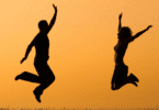 As silhuetas de um casal composto por uma mulher e um homem estão pulando no ar. Ao fundo, há o céu alaranjado do pôr do sol.