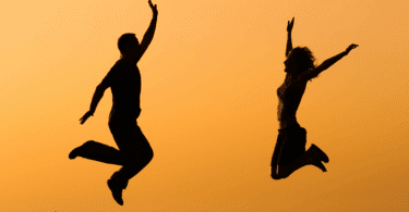 As silhuetas de um casal composto por uma mulher e um homem estão pulando no ar. Ao fundo, há o céu alaranjado do pôr do sol.