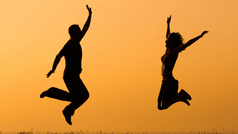 As silhuetas de um casal composto por uma mulher e um homem estão pulando no ar. Ao fundo, há o céu alaranjado do pôr do sol.