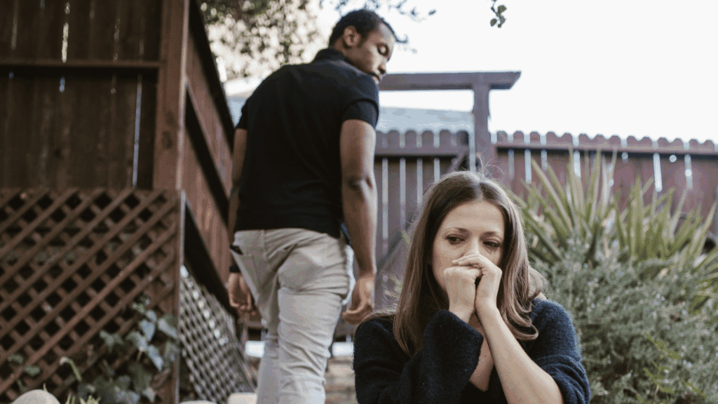 Mulher sentada ao ar livre com expressão triste e preocupada enquanto um homem se afasta caminhando, sugerindo um momento de tensão emocional.