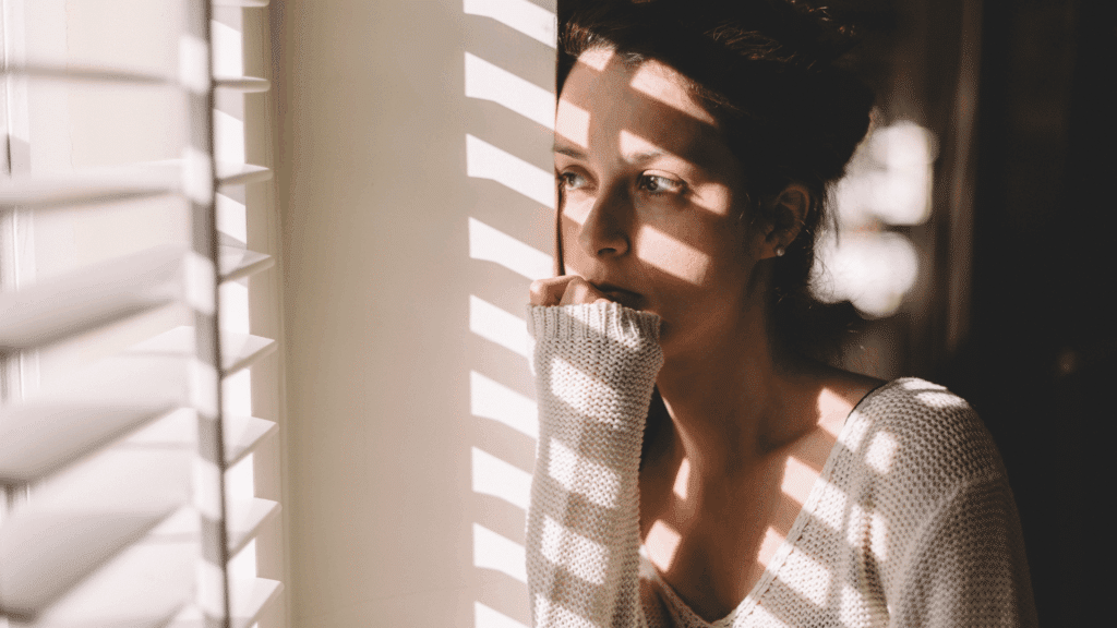 Mulher pensativa olhando pela janela, com luz entrando pelas persianas e criando sombras sobre seu rosto e suéter claro.