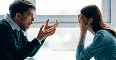 Homem gritando com as mãos erguidas enquanto uma mulher à sua frente cobre o rosto com as mãos, aparentando estar angustiada, em um ambiente interno iluminado por uma janela ao fundo.