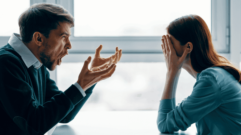 Homem gritando com as mãos erguidas enquanto uma mulher à sua frente cobre o rosto com as mãos, aparentando estar angustiada, em um ambiente interno iluminado por uma janela ao fundo.