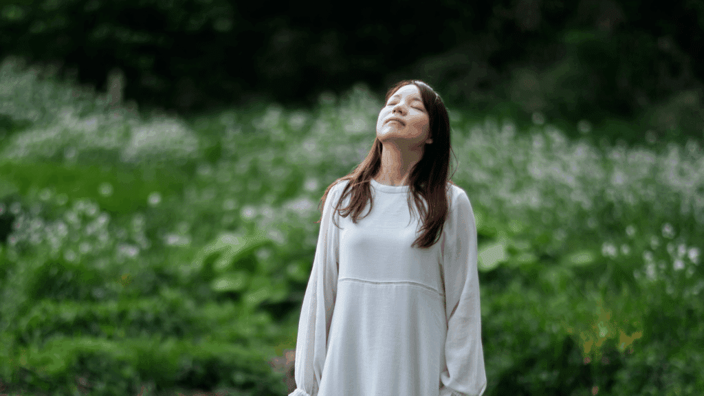 Uma mulher jovem de vestido branco está em um ambiente de natureza. Ela está com os olhos fechados e levanta a cabeça, respirando fundo e sentindo a atmosfera do lugar.