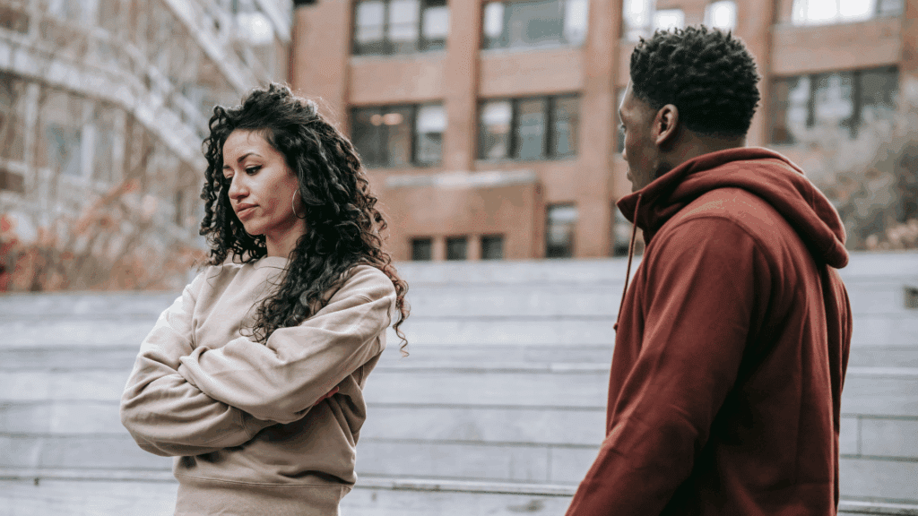 Mulher com os braços cruzados olhando para o lado com expressão de descontentamento enquanto um homem ao seu lado, vestido com moletom vermelho, parece tentar conversar com ela em um ambiente urbano ao ar livre.