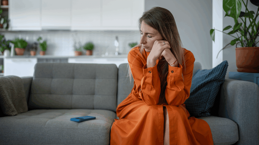 Mulher sentada em um sofá, com expressão triste e pensativa, olhando para um celular azul apoiado ao seu lado.