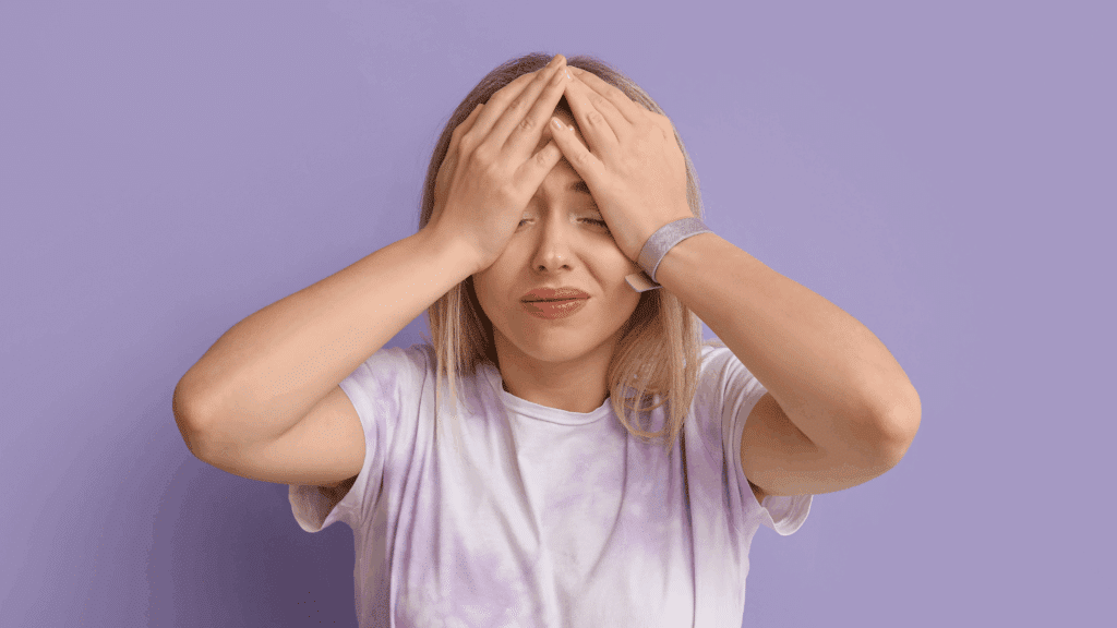 Mulher com expressão de frustração, com as mãos na cabeça e os olhos fechados, em frente a um fundo lilás.