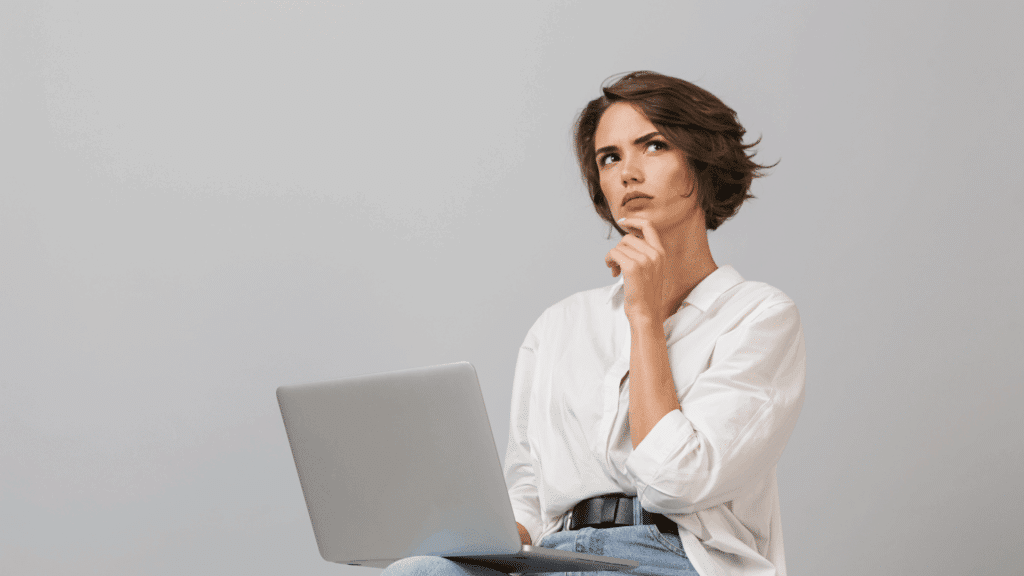 Uma mulher está sentada com o seu notebook aberto no colo. Ela coloca uma mão no rosto e olha para cima com uma expressão de dúvida, questionamento ou reflexão sobre algo.