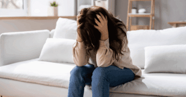 Mulher sentada no sofá com a cabeça abaixada e as mãos segurando o cabelo, aparentando estresse ou angústia.