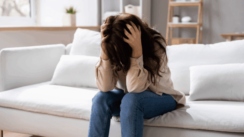 Mulher sentada no sofá com a cabeça abaixada e as mãos segurando o cabelo, aparentando estresse ou angústia.