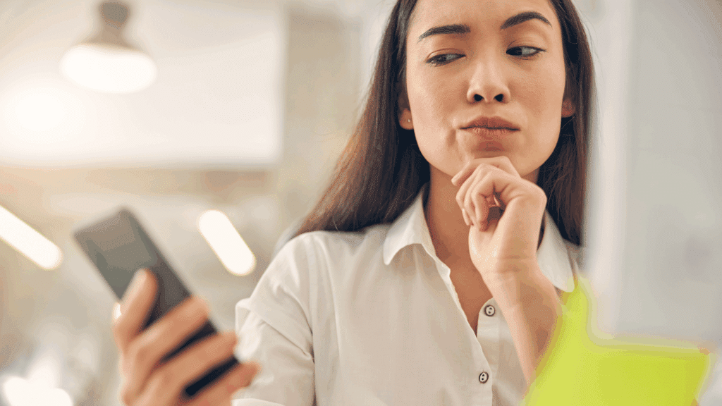Mulher olhando com desconfiança para o seu celular, o qual está na sua mão.