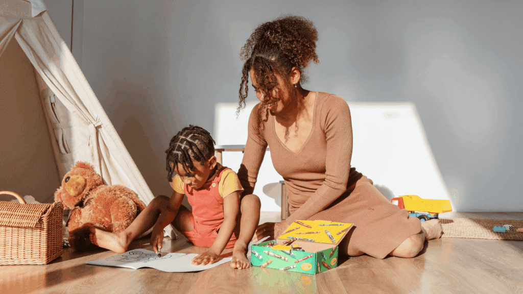 Uma mulher está brincando com seu filho. A criança está pintando em um papel ao lado dela.