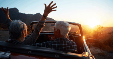 Um casal de idosos está dentro de um carro aberto (sem o teto). O carro está estacionado em um ambiente de natureza. Eles observam o pôr do sol e a mulher levanta os braços.