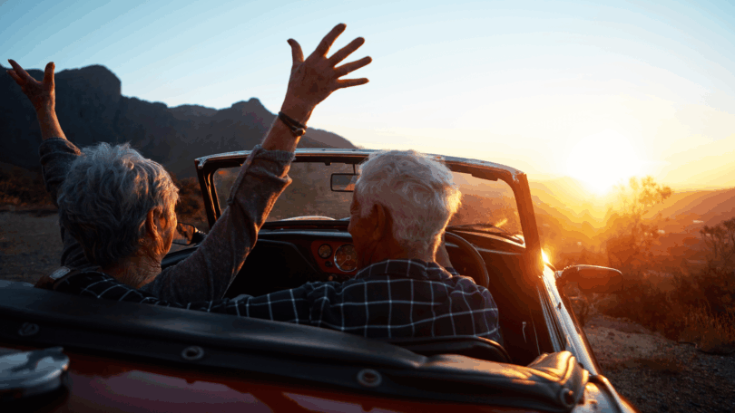 Um casal de idosos está dentro de um carro aberto (sem o teto). O carro está estacionado em um ambiente de natureza. Eles observam o pôr do sol e a mulher levanta os braços.