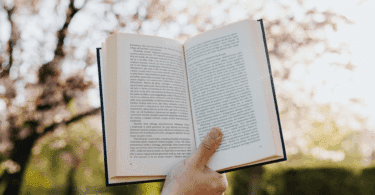 A mão de uma pessoa segura e levanta um livro aberto no ar. Ao fundo, há uma árvore e a luz do sol.