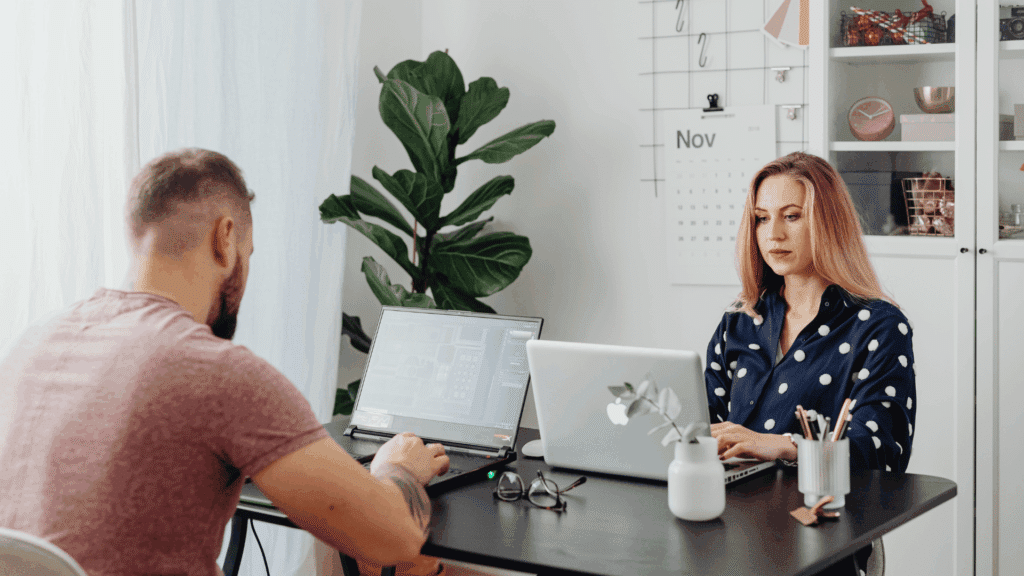Duas pessoas trabalhando em uma mesa: um homem de costas usando notebook à esquerda e uma mulher digitando em um laptop à direita, em um ambiente iluminado, com planta ao fundo e objetos de escritório sobre a mesa.