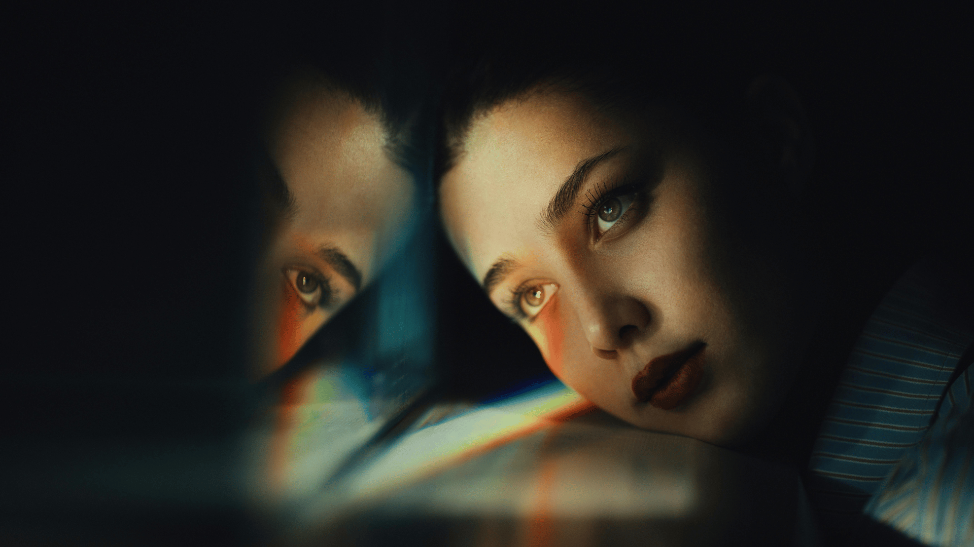 Mulher olhando pensativa para seu reflexo em uma superfície brilhante, iluminada por luz suave em ambiente escuro.