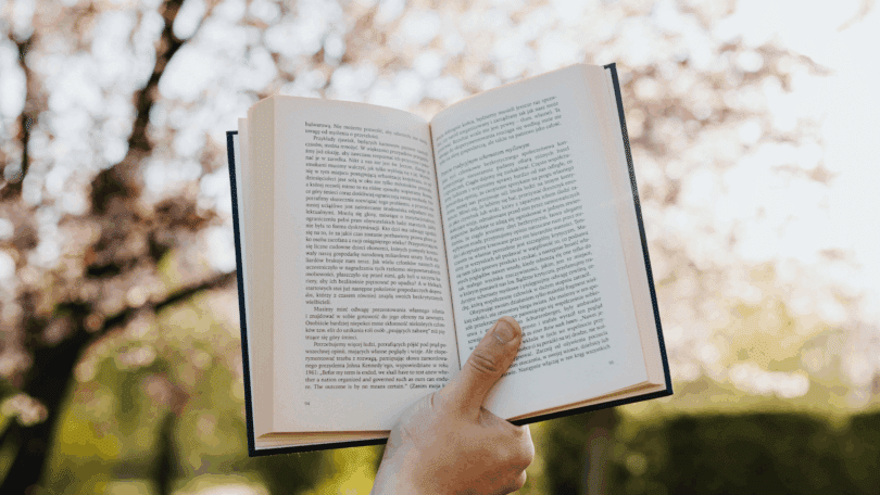 A mão de uma pessoa segura e levanta um livro aberto no ar. Ao fundo, há uma árvore e a luz do sol.