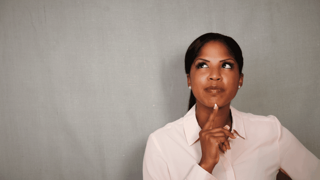 Mulher jovem olhando para cima com expressão pensativa, tocando o queixo, sobre fundo cinza.