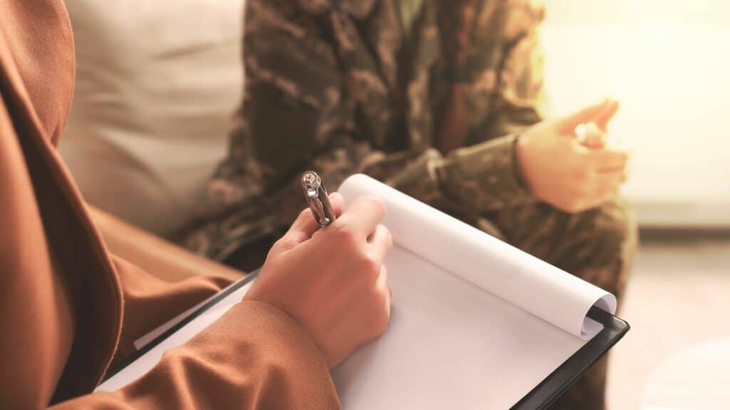 Imagem de uma pessoa fazendo uma consulta com um psicólogo. Eles estão sentados em um sofá e a profissional está fazendo anotações em um ficheiro de folhas brancas.