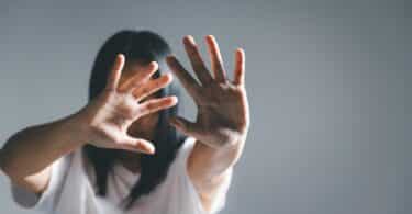 Imagem de uma mulher usando uma camiseta branca. Ela está com as mãos erguidas, protegendo o seu rosto, não querendo manter contato com ninguém, simbolizando o Ghosting que é o ato de interromper uma relação de forma abrupta e silenciosa.