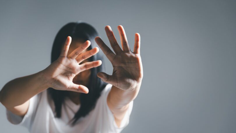 Imagem de uma mulher usando uma camiseta branca. Ela está com as mãos erguidas, protegendo o seu rosto, não querendo manter contato com ninguém, simbolizando o Ghosting que é o ato de interromper uma relação de forma abrupta e silenciosa.