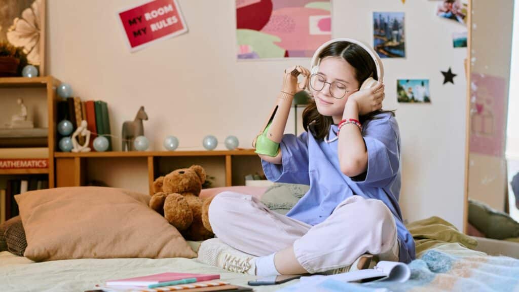 Imagem de uma adolescente em seu quarto sentada sobre a sua cama, ouvindo músicas com fones de ouvido.