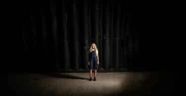 Imagem de uma mulher loira, usando uma jardineira jeans em cima de um palco e ao fundo, uma cortina preta, simbolizando a vida como um espetáculo.