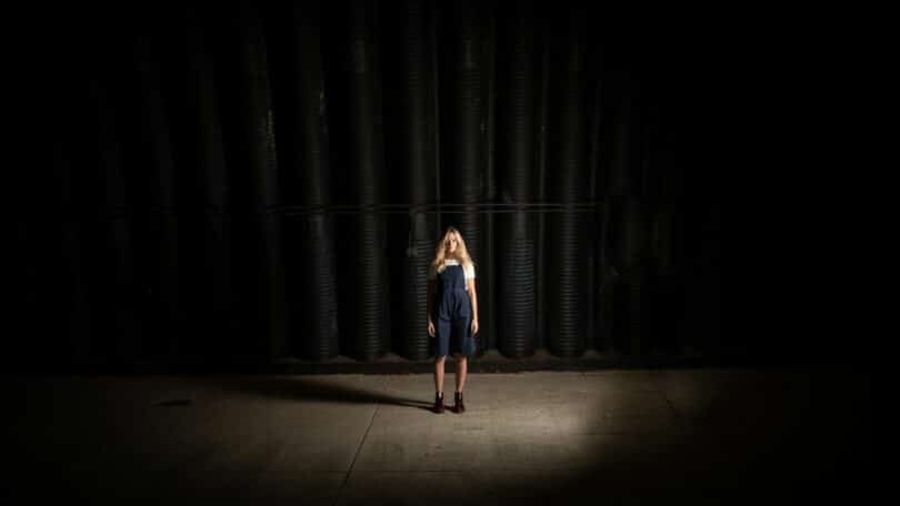 Imagem de uma mulher loira, usando uma jardineira jeans em cima de um palco e ao fundo, uma cortina preta, simbolizando a vida como um espetáculo.