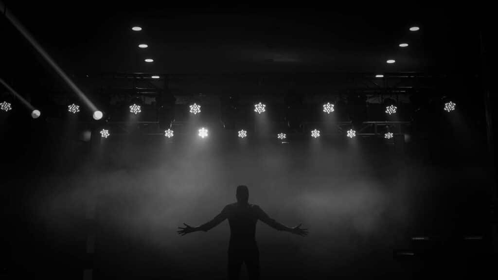 Imagem da silhueta de um homem sobre um palco com luzes branca no teto.