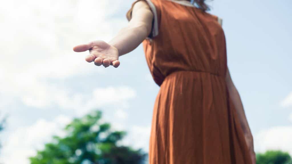 Imagem de uma mulher usando um vestido marrom, estendendo uma das mãos para ajudar uma pessoa, trazendo o conceito de pessoa bondosa.