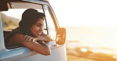 Imagem de uma mulher olhando o horizonte no meio de uma praia. Ela está dentro de uma kombi, contemplando o merecimento e a prosperidade da vida.