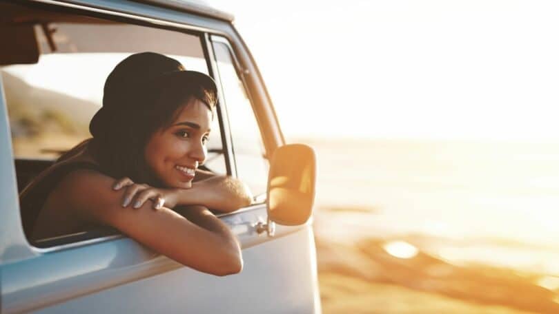 Imagem de uma mulher olhando o horizonte no meio de uma praia. Ela está dentro de uma kombi, contemplando o merecimento e a prosperidade da vida.