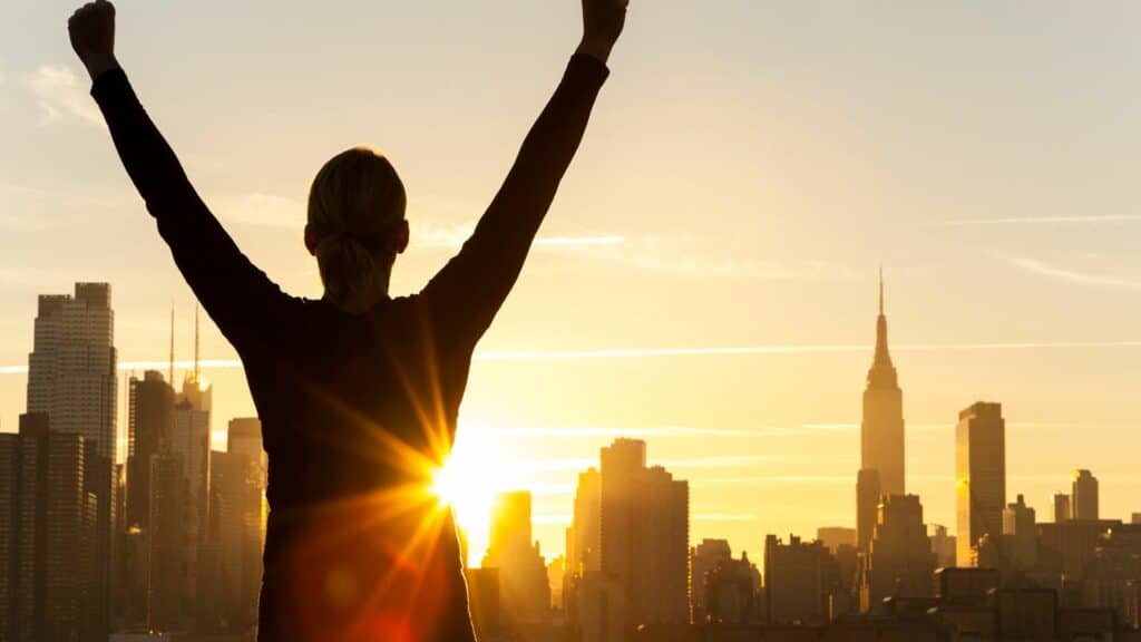Imagem da silhueta de uma pessoa olhando para o horizonte cheio de prédios e um lindo pôr do Sol, simbolizando o conceito de estar ocupando o seu lugar no mundo sem pedir licença e a reconhecer seu valor sem precisar provar nada.