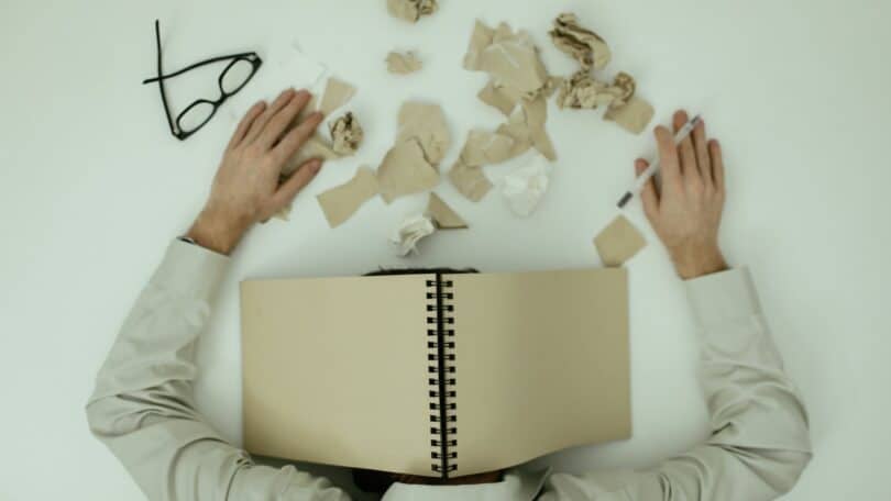 Imagem de uma pessoa debruçada sobre uma mesa, com o óculos de grau deixado ao lado, além de vários papéis picados. A foto simboliza uma pessoa com os sintomas do Transtorno da Sobrecarga Sensorial.