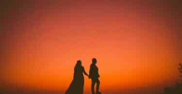 Imagem de fundo alaranjado e em destaque um casal de mãos dadas olhando para o pôr do Sol, trazendo o conceito de amor secreto.