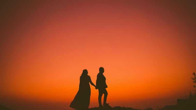 Imagem de fundo alaranjado e em destaque um casal de mãos dadas olhando para o pôr do Sol, trazendo o conceito de amor secreto.