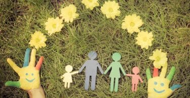 Imagem das mãos de uma criança sobre um gramado contendo figuras em papel de um homem, de uma mulher e de duas crianças, rodeadas com flores amarelas, representando o universo familiar.