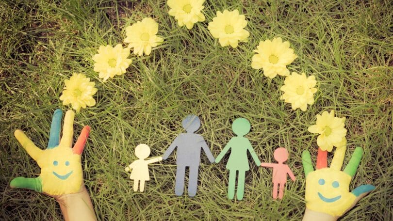 Imagem das mãos de uma criança sobre um gramado contendo figuras em papel de um homem, de uma mulher e de duas crianças, rodeadas com flores amarelas, representando o universo familiar.