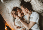 Casal deitado abraçado em uma cama, sorrindo e encostando os rostos com carinho, em um ambiente acolhedor com almofadas texturizadas ao fundo.