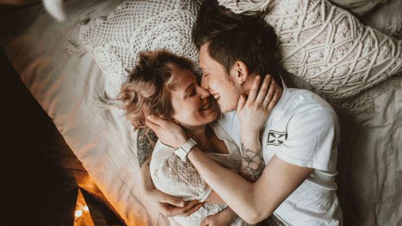 Casal deitado abraçado em uma cama, sorrindo e encostando os rostos com carinho, em um ambiente acolhedor com almofadas texturizadas ao fundo.