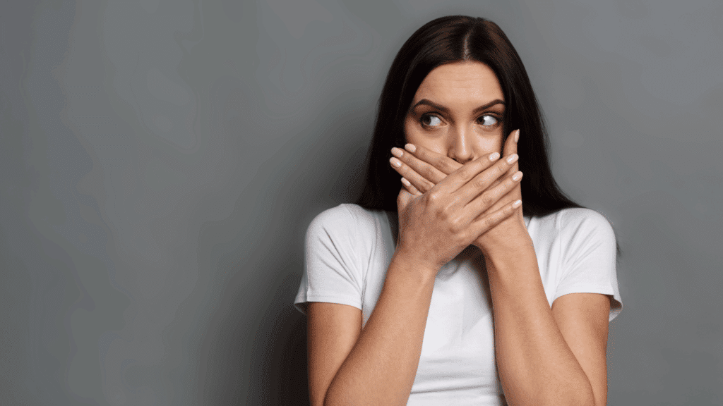 Um mulher jovem que veste uma t-shirt branca está em destaque na imagem. Ela está com as duas mãos cobrindo a boca e olha para o lado, indicando não poder falar algo, ou ter falado algo que não deveria.