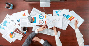 Os braços de dois funcionários de uma empresa segurando papéis com resultados e informações sobre negócios estão em destaque na imagem. Em cima de uma grande mesa, há diversos papéis com dados, há um computador, um vaso com planta, uma agenda, uma caneta, uma calculadora e um celular. Os funcionários conversam sobre os dados.