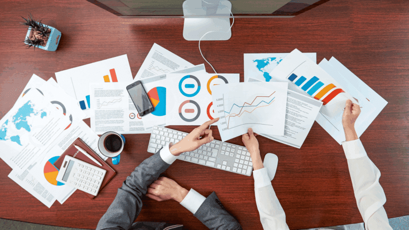 Os braços de dois funcionários de uma empresa segurando papéis com resultados e informações sobre negócios estão em destaque na imagem. Em cima de uma grande mesa, há diversos papéis com dados, há um computador, um vaso com planta, uma agenda, uma caneta, uma calculadora e um celular. Os funcionários conversam sobre os dados.