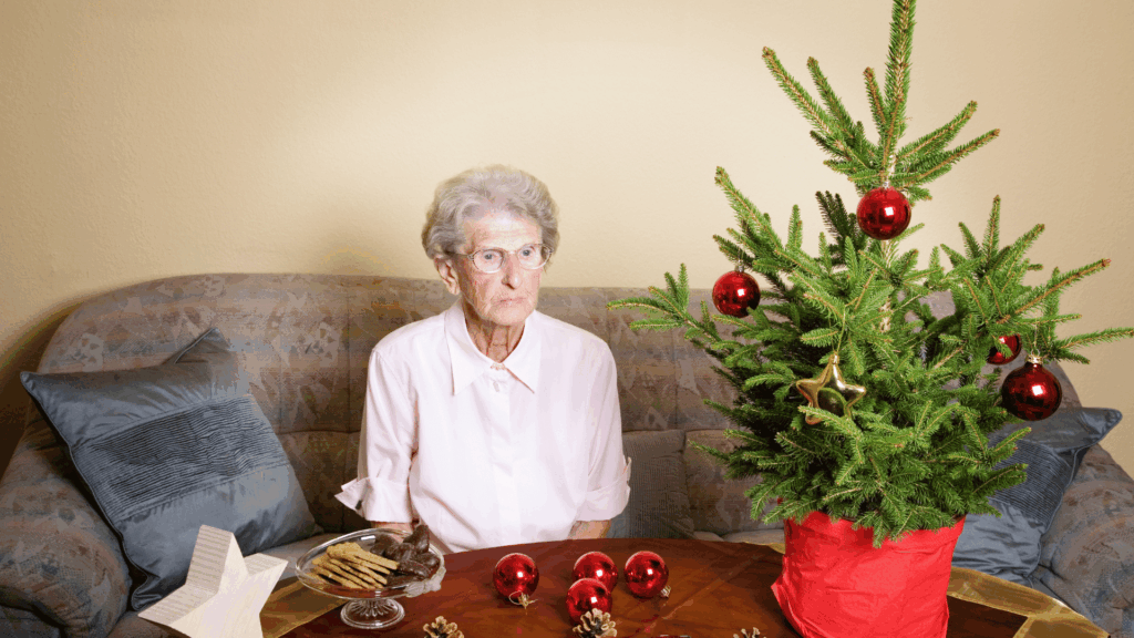 Um mulher idosa está sentada de frente para uma mesa. É época de Natal. Na mesa, há decorações de Natal, uma árvore de Natal e chocolates. A senhora está com uma feição no rosto de desamparo, tristeza.