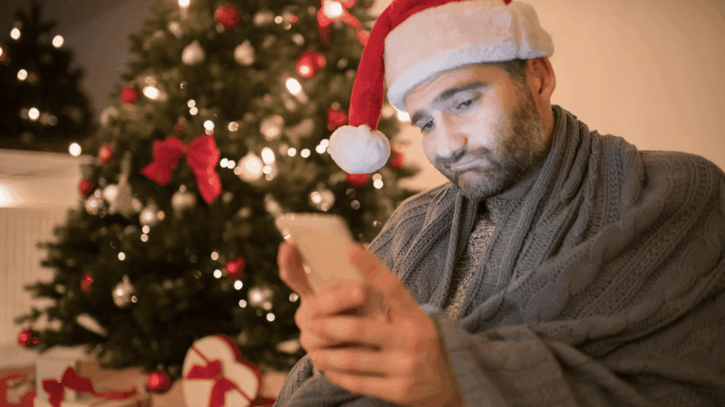 Um homem está mexendo no seu celular e apresenta uma feição de tristeza no rosto. É época de Natal e há uma árvore de Natal ao fundo.