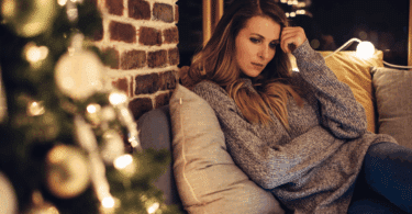 Uma mulher jovem está sentada em um sofá. Ela está com uma mão na cabeça e uma feição triste no rosto. É época de Natal e há uma árvore de Natal ao lado.