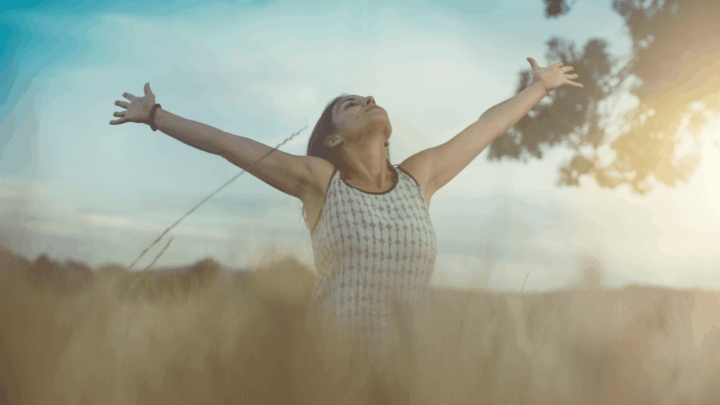 Uma mulher jovem está em um ambiente aberto de natureza. Ela levanta a cabeça e os braços para o ar e sente a brisa do lugar.