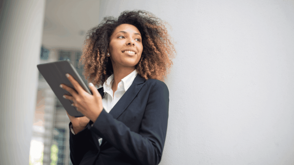Um mulher jovem e de negócios está sorrindo e olhando para o lado. Ela segura um tablet nas mãos.